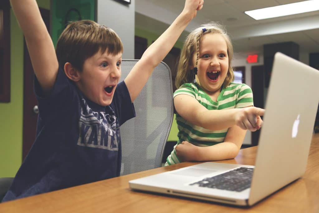 This is a picture of a little girl excitedly pointing at a computer screen. Next to a little boy with his arms up  